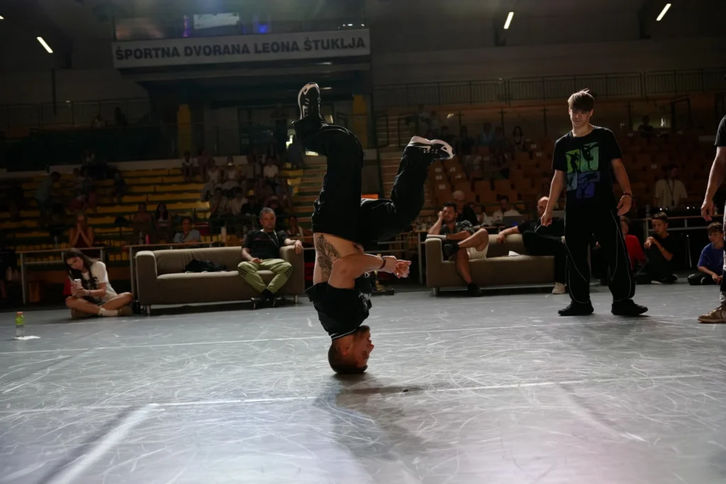 Breakdance tekmovanje – plesalec Bast Arts izvaja headspin pred občinstvom v dvorani.